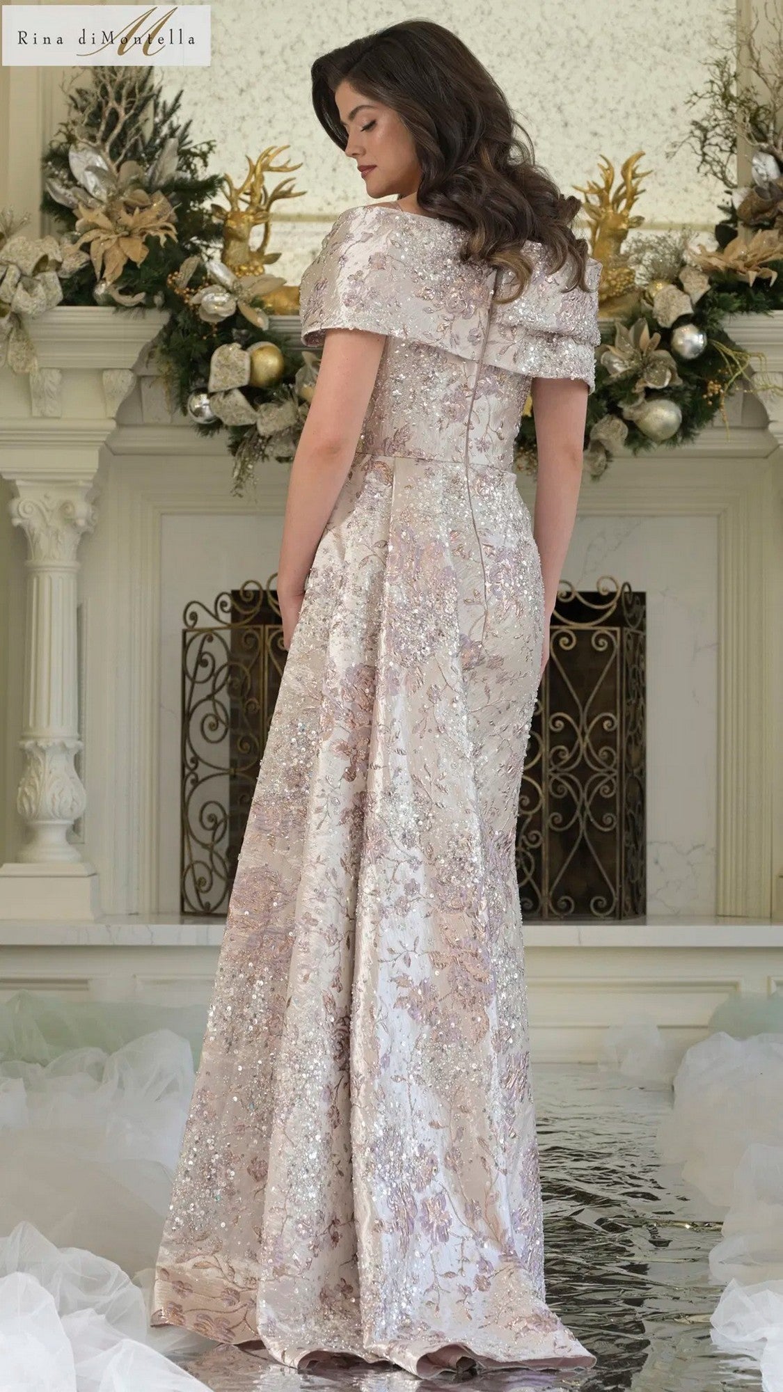 A woman stands indoors in a Rina Di Montella RD3306 long formal dress featuring an off-the-shoulder jacquard design with a shimmering floral pattern. Decorative greenery and gold ornaments are visible in the background.